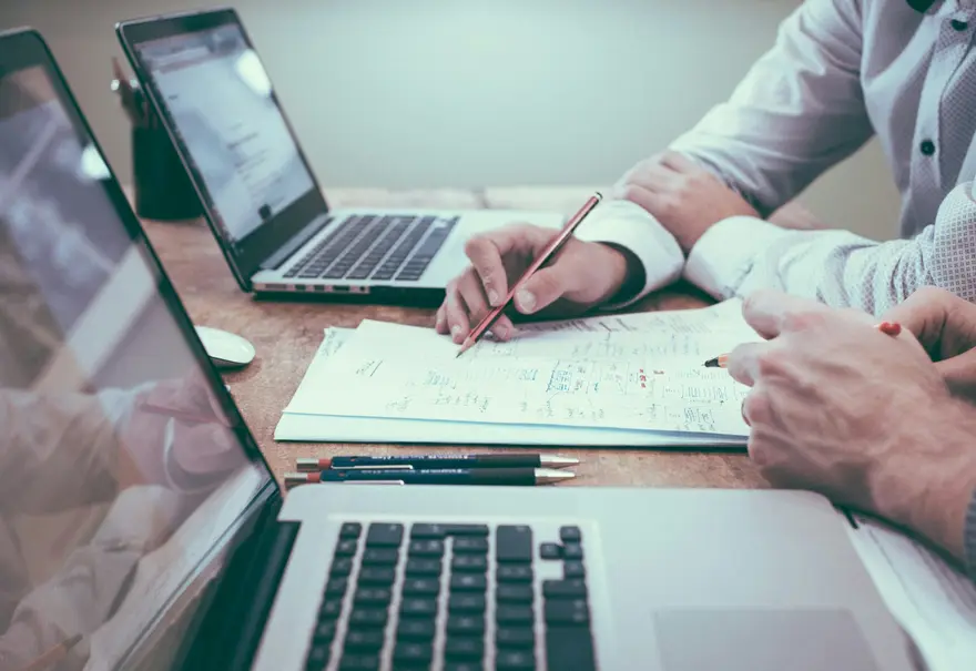 Dos personas revisando un cuaderno junto a portátiles abiertos sobre una mesa de trabajo